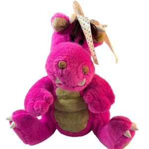 May include: A plush dragon toy in vibrant fuchsia with a textured tan belly and light-colored claws on its feet. The creature features dark brown hair on its head, adorned with a white ribbon decorated with colorful polka dots tied in a bow.