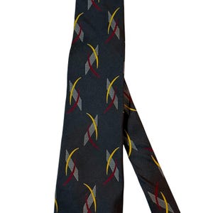 May include: A dark navy blue necktie featuring a repeating pattern of abstract geometric shapes. Each motif consists of a grey hatched rectangle overlaid with a curved yellow line and a curved dark red line, creating a consistent graphic design.