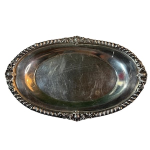 Puede incluir: Bandeja ovalada plateada con detalles florales y de volutas en relieve en el borde. La superficie de la bandeja presenta algunos arañazos. La forma general es alargada, con un aspecto brillante y decorativo.
