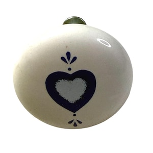 May include: A round, off-white ceramic cabinet knob featuring a dark blue heart design centered on the front. The heart is flanked by small, stylized floral motifs above and below it. A metallic mounting hardware piece is visible at the top.