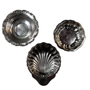 May include: Three antique silver serving bowls. The bowls have intricate designs, including a scalloped edge and a shell-shaped bowl. The bowls are arranged on a white background, showcasing their reflective surfaces.