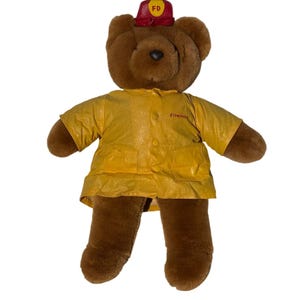 May include: A brown teddy bear wearing a yellow fireman's jacket and a red firefighter's hat. The hat has the letters "FD" on it. The jacket has the word "fireman" printed on it.