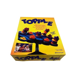 May include: A vintage "Topple" board game box with a yellow background and the game's name in large blue letters. The game features colorful stacked discs on a blue platform. The box includes text that reads "Winning or losing hangs in the balance of every move!"