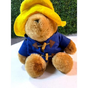 May include: A plush teddy bear wearing a bright yellow hat and a blue coat with wooden toggle closures. The bear has brown fur and is sitting down. The coat has brown leather-like patches.