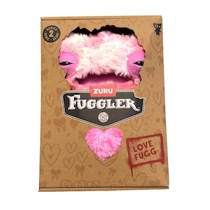 Op de afbeelding: Een roze en wit Fuggler pluche speelgoed komt uit een kartonnen doos. De doos heeft het Fuggler-logo, een roze hart en de woorden "LOVE FUGG". Het speelgoed heeft grote, donkere ogen en een gestikte mond.