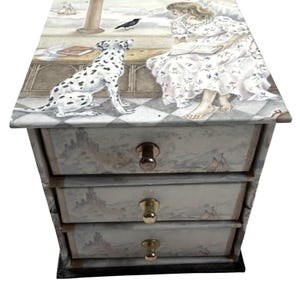 May include: A small wooden chest of drawers featuring three drawers with gold-toned knobs. The exterior is decorated with a printed illustration of a person reading a book, a dalmatian dog, a black bird, and a coastal landscape with a castle.