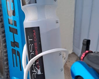 May include: A translucent white plastic water bottle with a black cap and a black label featuring the text GHOST BIKES.COM. The bottle is held in a white cage mounted on a bright blue bicycle frame, with visible volume markings at 125ml, 250ml, and 500ml.