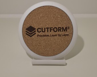 May include: A round coaster with a white plastic frame and a cork center. The cork features a dark brown logo with a stacked square icon, the text CUTFORM3, and the tagline Precision. Layer by Layer. The coaster rests on a small white stand.