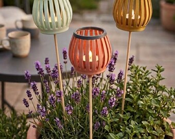 May include: Three garden stakes featuring spherical, slatted candle holders in light green, terracotta, and gold. Each stake holds a small, lit tea light candle and is positioned within a large terracotta planter filled with lavender and green herbs.