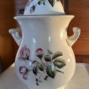 Puede incluir: Jarra de cerámica color crema con tapa, adornada con rosas rosas y hojas verdes. La jarra tiene dos asas ornamentadas y un ribete dorado. La tapa presenta un pequeño asa y un diseño floral. Una pieza decorativa para el hogar.