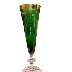 May include: A tall, green champagne flute with a gold rim and a decorative base. The glass has a slender, conical shape and is adorned with red accents. The base features a white surface with red and green decorative lines.