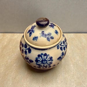 Puede incluir: Un pequeño tarro redondo de cerámica con tapa. El tarro es de color blanquecino con diseños florales azules y un pomo marrón oscuro en la tapa. El diseño floral incluye flores y hojas estilizadas. El tarro es probablemente para guardar objetos pequeños.