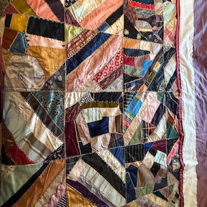 May include: A colorful patchwork quilt with a variety of fabrics and patterns. The quilt features diagonal stripes and geometric shapes in shades of pink, blue, black, and gold. The edges are finished with a white border and a pink ribbon.