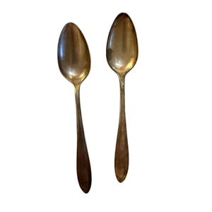 May include: Two metallic spoons with a tarnished silver-toned finish. Each spoon features an oval-shaped bowl and a smooth, plain handle that tapers toward the end. The spoons are displayed vertically, showing a consistent design and color.