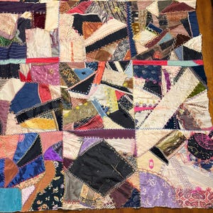 Könnte beinhalten: Ein Patchwork-Quilt mit einer Vielzahl von Stoffstücken in verschiedenen Farben und Mustern. Der Quilt zeigt Quadrate und Dreiecke aus verschiedenen Texturen, darunter Samt, Satin und gemusterte Baumwolle. Die Kanten sind mit einem Kontrastfaden genäht.