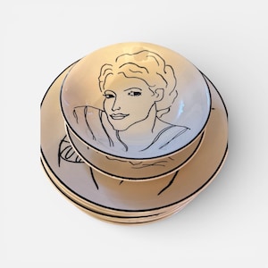 May include: A stack of beige ceramic plates and a bowl featuring a black outline drawing of a woman's face. The plates and bowl have a black trim. The woman's face is drawn in a minimalist style.