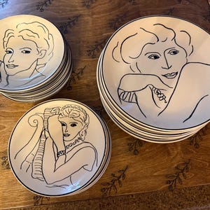 May include: A set of white ceramic plates featuring black line art portraits of women. Each plate has a black rim. The plates are stacked in three piles of different sizes. They are arranged on a wooden surface.