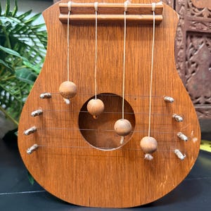 Puede incluir: Un instrumento de cuerda de madera con un cuerpo redondeado y un agujero de sonido central. El instrumento tiene cuatro cuerdas y cuentas de madera colgando de ellas. La madera tiene un tono marrón cálido.