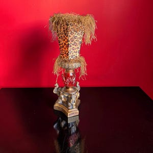Può includere: Un portacandele decorativo con paralume con stampa leopardata e centro in vetro rosso. La base è color oro con dettagli di elefanti. Il paralume è rifinito con una frangia di materiale simile alla paglia.