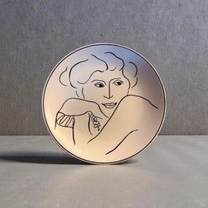 May include: A white ceramic plate with a black outline drawing of a woman's face and upper body. The artwork is in a minimalist style, with the woman's face and form depicted in simple, elegant lines. The plate has a black border.