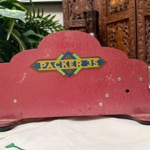 Op de afbeelding: Een vintage rode Packer 35 tapedispenser met een decoratief label. De dispenser heeft een gebogen bovenkant en een metalen snijkant. Het label toont de tekst "PACKER 35" in een ruitvormig ontwerp.