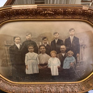 Puede incluir: Fotografía antigua enmarcada en un marco ovalado dorado. La imagen en sepia muestra un retrato familiar. Los miembros, adultos y niños, visten ropa formal. El marco es ornamentado.