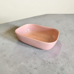 May include: A small, rectangular, pink ceramic dish with rounded corners. The interior has a slightly lighter shade of pink. The dish is empty and sits on a gray surface.