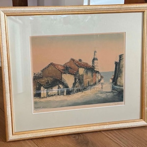May include: A framed print featuring a landscape illustration of houses with red roofs and a tall tower under a soft peach-colored sky. The artwork is enclosed in a light-colored mat and a gold-toned frame with a textured, decorative border finish.
