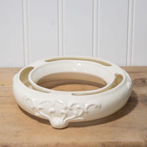 May include: A cream-colored, ceramic centerpiece bowl with a circular design. The bowl features an inner ring and decorative floral relief patterns. It sits on a wooden surface against a white paneled background.