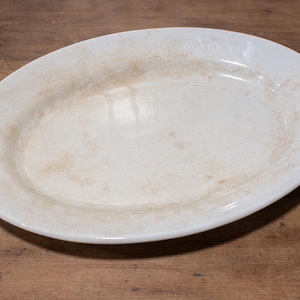 May include: An oval-shaped white ceramic serving platter featuring a smooth, glossy finish with subtle, light brown mottled staining or discoloration distributed across the surface, creating a weathered appearance on the otherwise plain white dish.