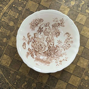 May include: A white ceramic bowl with a scalloped edge, featuring a brown floral design. The design includes a basket of flowers and other floral elements. The bowl is set on a gold and black checkered surface.