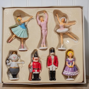 May include: A set of six Nutcracker-themed ornaments in a plastic tray. The figures include three dancers in blue, pink, and white costumes, a mouse king with a sword, a soldier in a red uniform with a rifle, and a girl in a purple dress holding a candle.