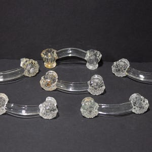 May include: A set of five clear glass drawer pulls, each featuring a curved handle and two faceted, star-shaped mounting ends. The transparent glass components have a smooth, arched center section and textured, multi-faceted circular bases.