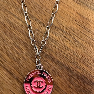 May include: A silver-toned metal chain necklace featuring a circular pendant with a pink enamel inlay. The pendant displays the text CHANEL PARIS 31 RUE CAMBON and a central interlocking CC logo surrounded by a serrated edge design.