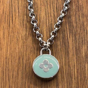 May include: Silver-toned necklace with a round, light blue pendant. The pendant features a silver flower design in the center and is attached to a chain. The background is a wooden surface.