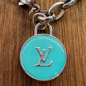 May include: A silver-toned metal chain necklace featuring a circular pendant with a bright turquoise enamel inlay. The center of the pendant displays a raised silver-colored LV monogram logo. The pendant is attached to the chain by a small silver loop.