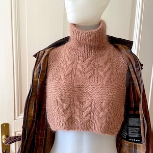 May include: A cropped, cable-knit turtleneck sweater in a dusty rose hue. The sweater has a high collar and a textured design. It is displayed on a mannequin, with a brown jacket visible in the background.