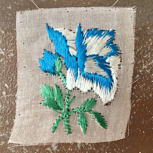 May include: A white fabric square with a blue and white embroidered flower with green leaves.