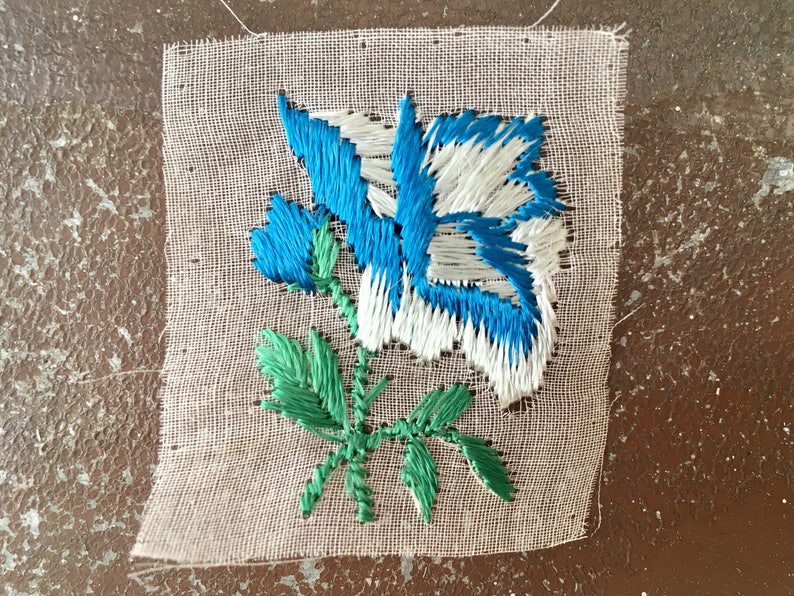 May include: A white fabric square with a blue and white embroidered flower with green leaves.