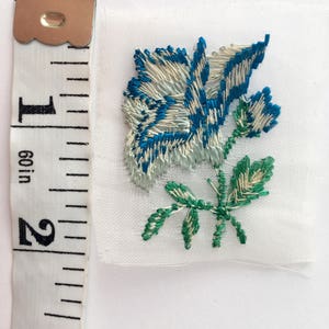 May include: A white fabric applique with a blue and white embroidered rose and green leaves.