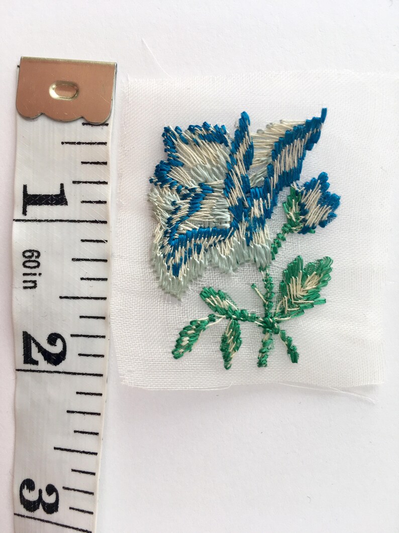 May include: A white fabric applique with a blue and white embroidered rose and green leaves.