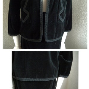 May include: A black velvet suit with a trim of gray piping. The jacket has a collar and long sleeves. The skirt is knee-length.