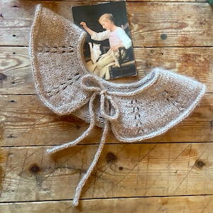 Könnte beinhalten: Ein hellbrauner, handgestrickter Kragen mit zartem, gewelltem Rand und Schleifenverschluss. Der Kragen ist aus weichem Garn gefertigt und hat eine Vintage-Ästhetik. Eine Vintage-Postkarte mit einer Frau und einer Katze befindet sich im Hintergrund.