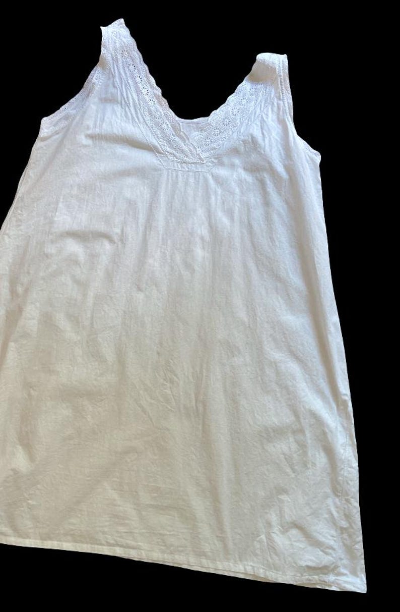 May include: A white sleeveless nightgown with a V-neckline and delicate lace trim. The fabric appears to be lightweight and slightly textured. The garment is likely vintage or inspired by vintage styles.