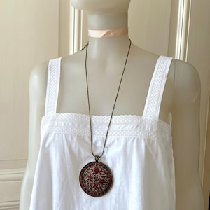 May include: A long, thin, silver chain necklace with a large, round pendant. The pendant is made of metal and has a red, floral design.