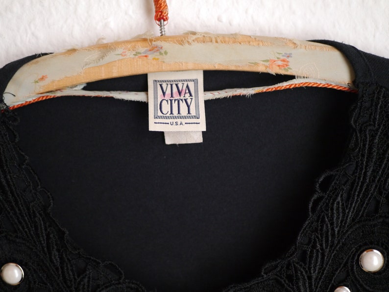 Może przedstawiać: Czarny top z misternymi koronkowymi detalami i perłowymi zdobieniami. Etykieta głosi "VIVA CITY USA". Top wisi na drewnianym wieszaku w stylu vintage z kwiatowymi akcentami.