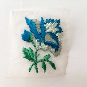 May include: A white fabric patch with a blue and white embroidered rose and green leaves.