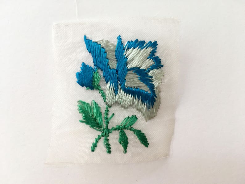 May include: A white fabric patch with a blue and white embroidered rose and green leaves.