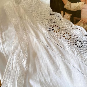 May include: White linen fabric with intricate lace detailing. The lace features floral and geometric patterns along the edges. A button is visible near the top. The fabric is folded, showing texture and depth. The background includes a painting.