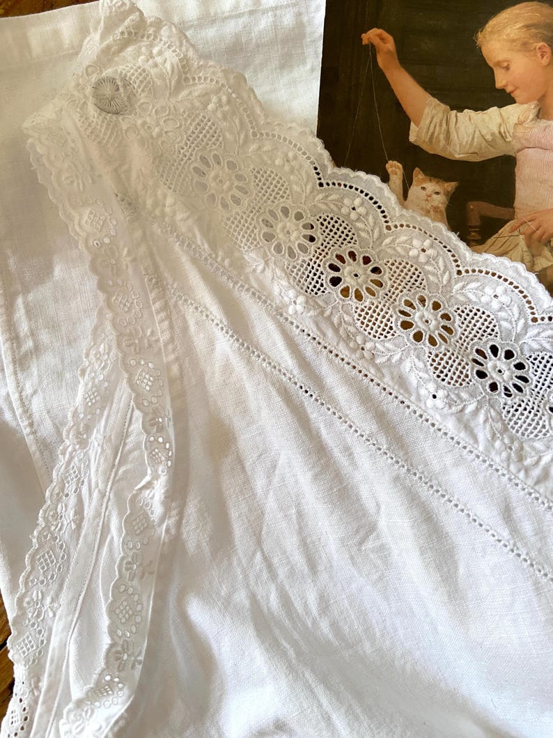 May include: White linen fabric with intricate lace detailing. The lace features floral and geometric patterns along the edges. A button is visible near the top. The fabric is folded, showing texture and depth. The background includes a painting.